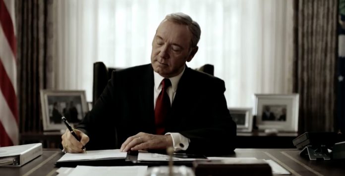 Il presidente Underwood è seduto alla scrivania dello studio ovale. Indossa un completo nero e una cravatta rossa. L'espressione è quella della soddisfazione, mentre firma un editto con una postura piena di sé. E' uno dei tratti distintivi del protagonista di House of Cards