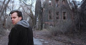 Un biondo protagonista ci da la schiena, ma il suo sguardo si gira sopra la sua spalla, insospettito. Sullo sfondo, alberi spogli e il rudere di una vecchia casa.