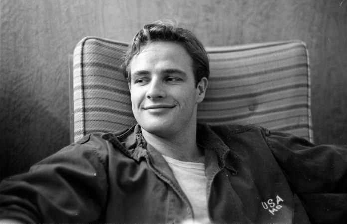 Hollywood sta preparando la biopic su Marlon Brando, qui ritatto da giovane, in mezzo busto, seduto su una poltrona, con un sorriso molto goduto.